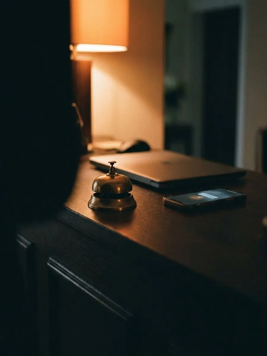 A hotel reception desk at dusk with a brass service bell, a laptop and a phone lighting up with a new booking